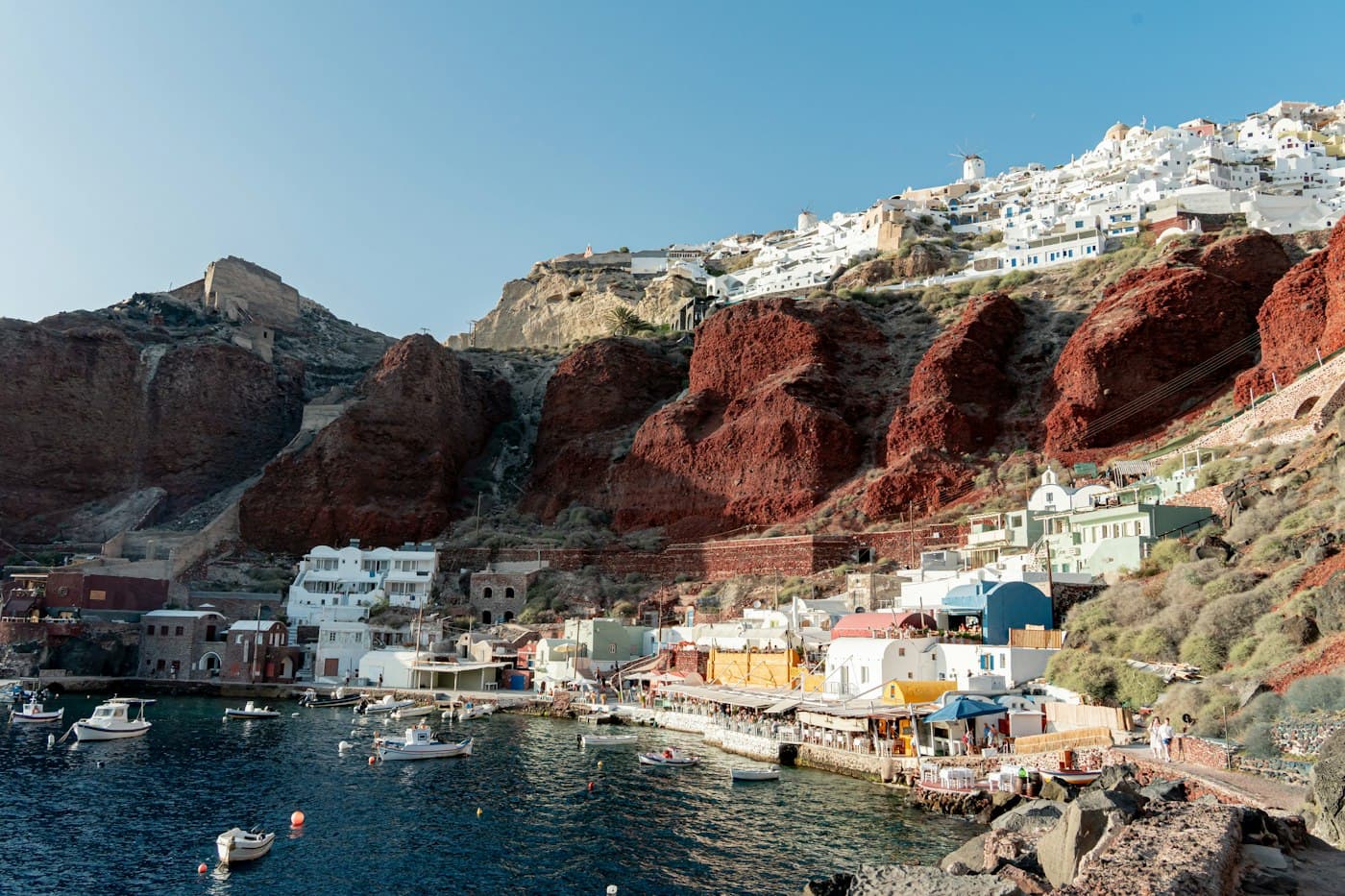Ammoudi Bay below Oia: small boats in the harbour, tavernas along the quay, red volcanic cliffs and Oia’s white houses and windmill on the rim above