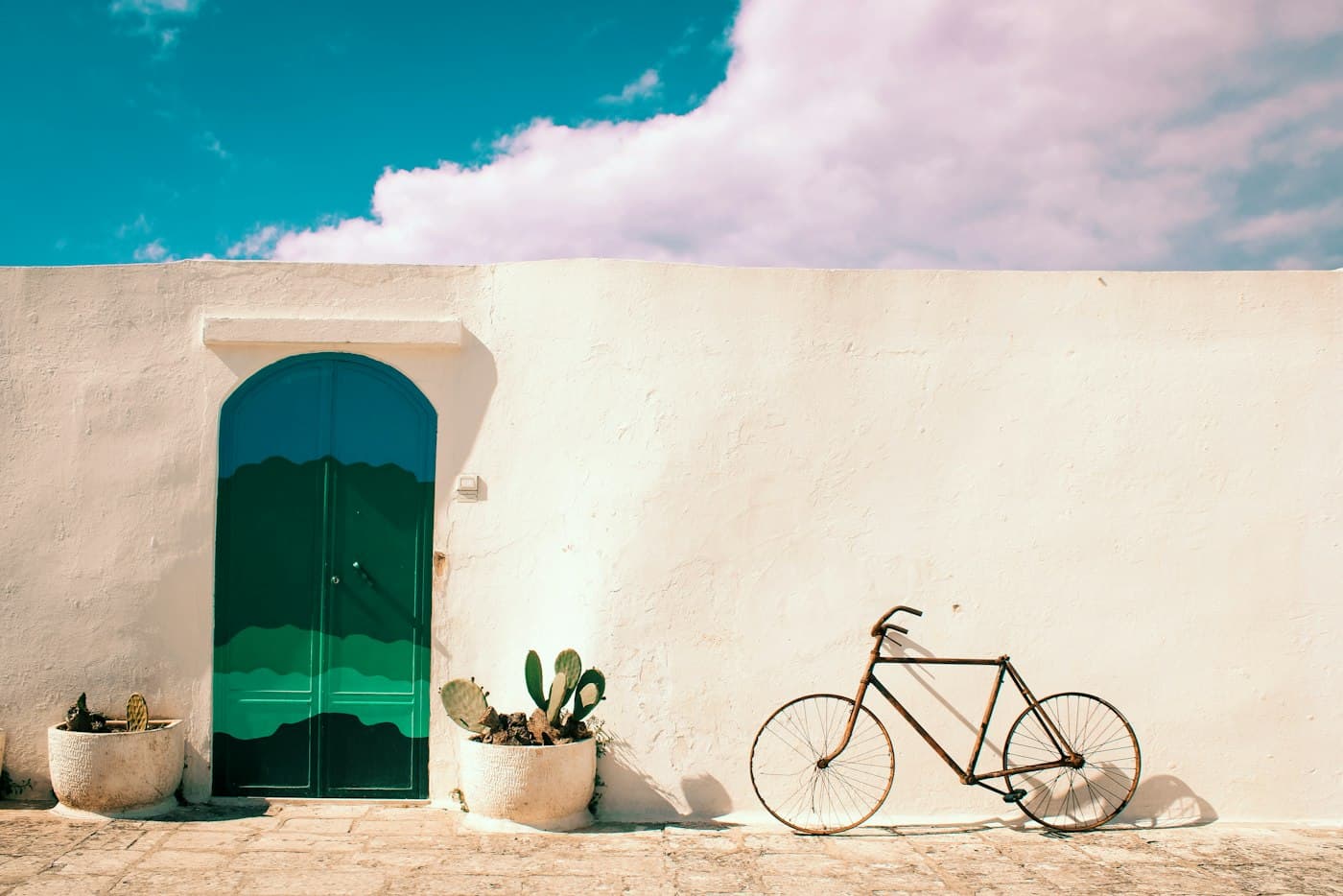 A green arched door, potted prickly pears and an old bicycle against a whitewashed wall in Ostuni