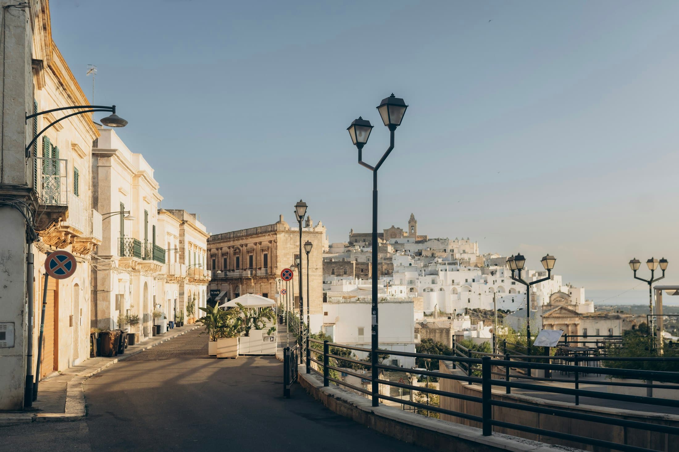A quiet street in Ostuni in low sun, lamp posts along the railing and the white old town on its hill beyond