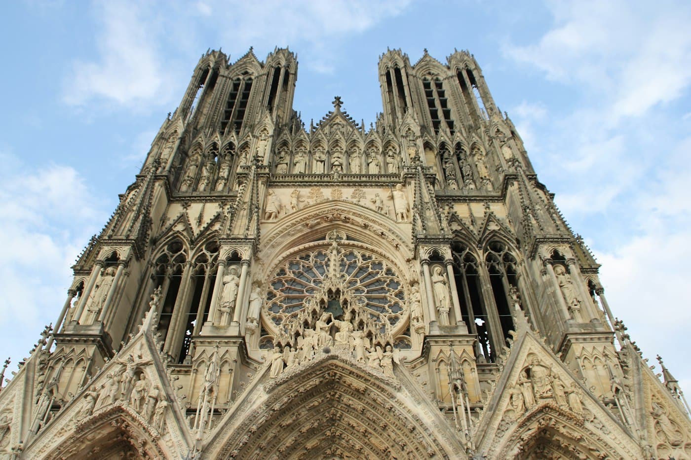 The west front of Reims Cathedral seen from below: its two towers, the great rose window and rows of carved figures against a pale sky