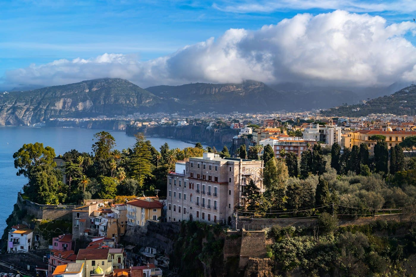 Vecchi alberghi e giardini sul bordo della falesia a Sorrento, pini e palme sopra il mare, la costa e le sue montagne sotto nuvole bianche alle spalle