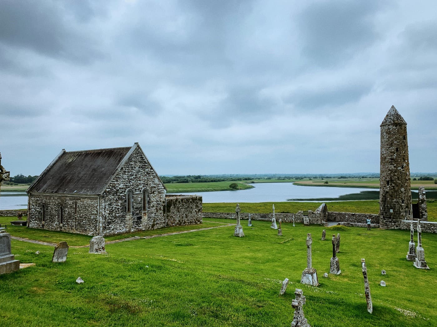 The monastic site of Clonmacnoise in County Offaly: a slate-roofed stone church, a round tower and old grave crosses on the grass, with the River Shannon winding through flat fields behind