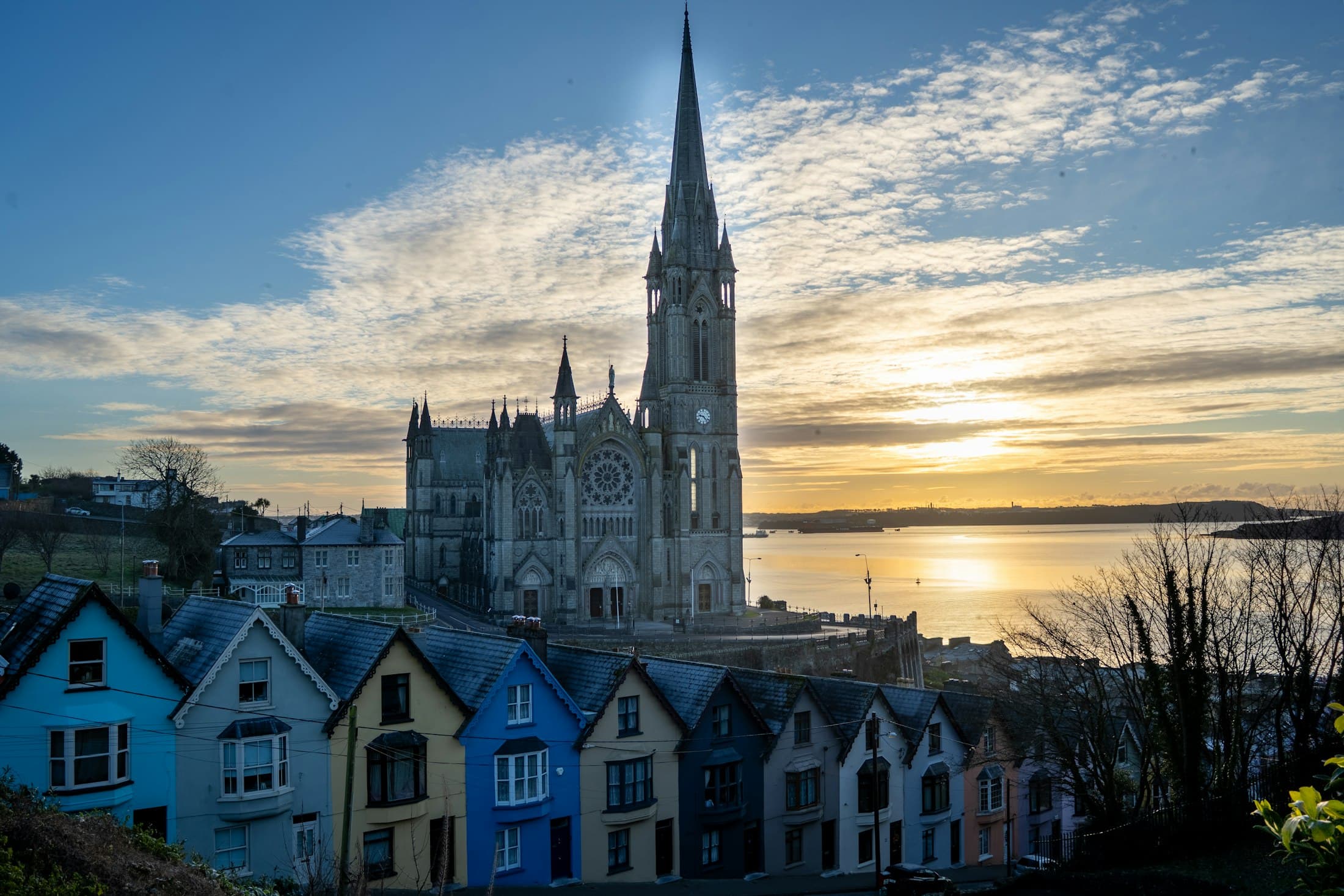 La cattedrale di San Colman sopra una fila di case colorate a Cobh all’alba, con il porto di Cork che luccica dietro