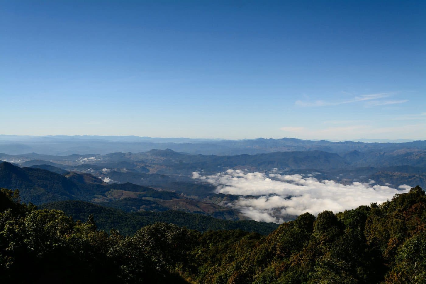Der Blick vom Doi Inthanon über bewaldete Hänge, ein Meer aus weißen Wolken im Tal und Kette um Kette blauer Berge unter klarem Himmel