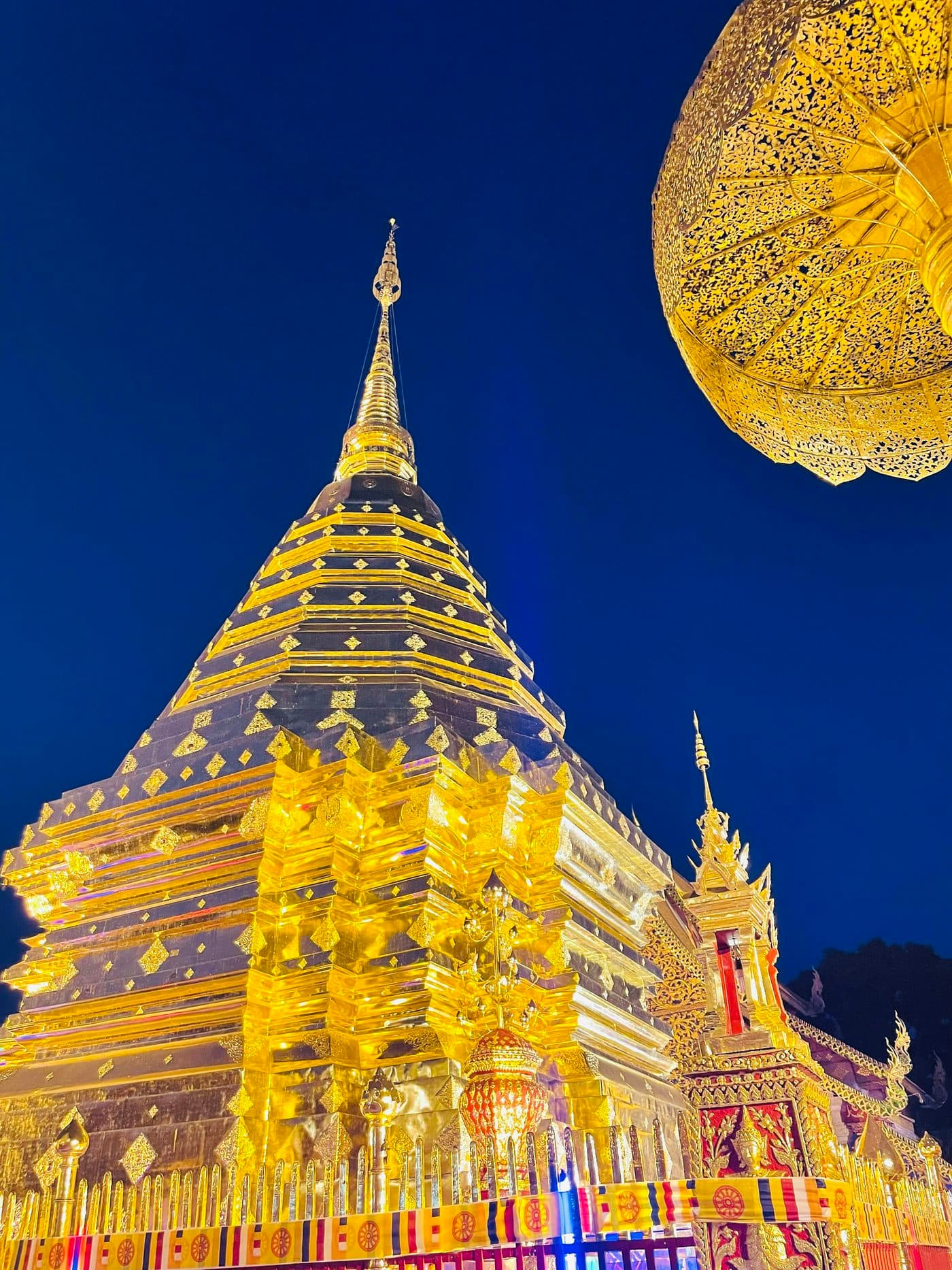 Der vergoldete Chedi des Wat Phra That Doi Suthep, nachts angestrahlt vor tiefblauem Himmel, an einer Ecke ein goldener Zeremonienschirm