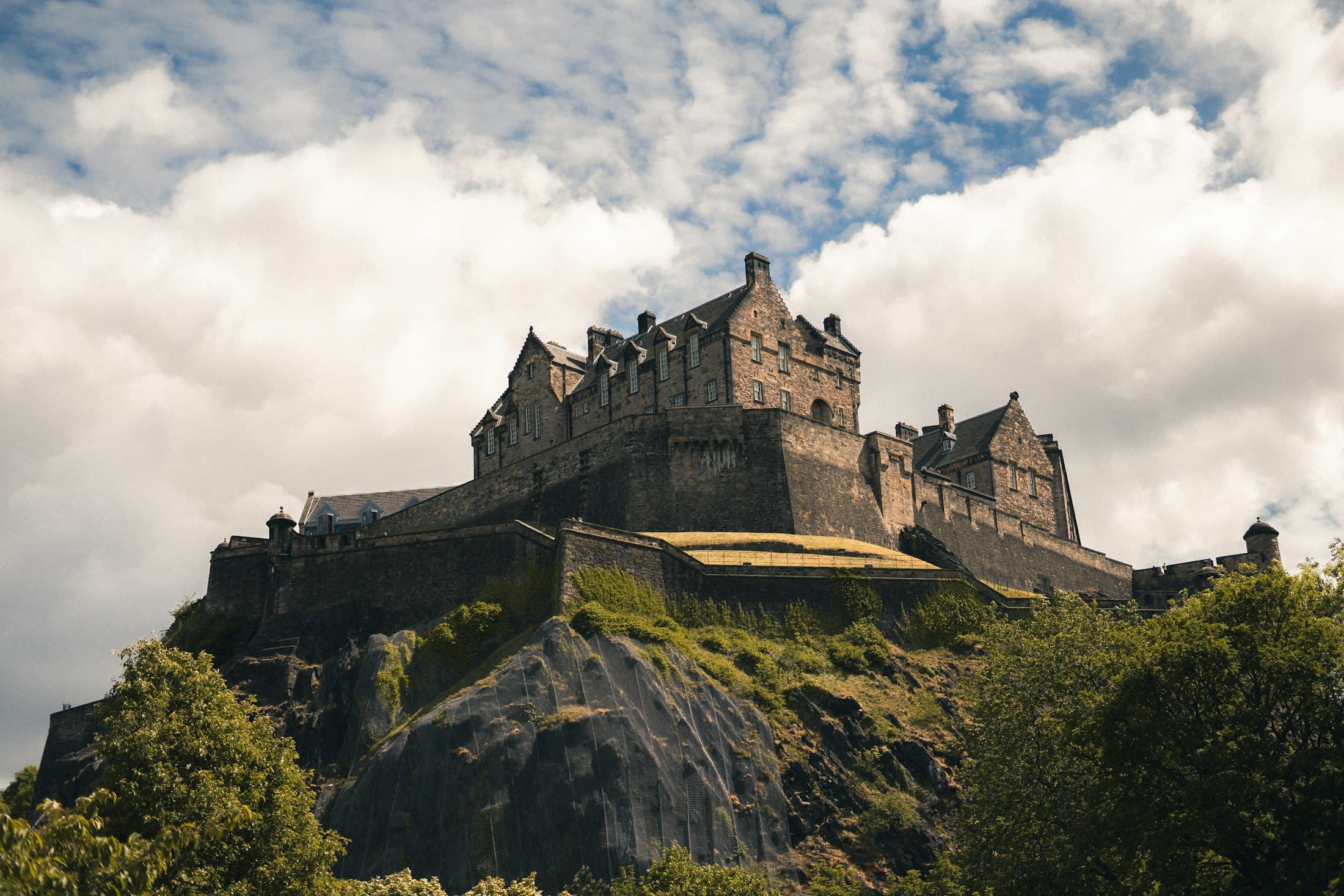Die Steinmauern und Giebelbauten von Edinburgh Castle auf seinem dunklen Vulkanfelsen über grünen Bäumen, unter weißen Wolken und blauen Lücken