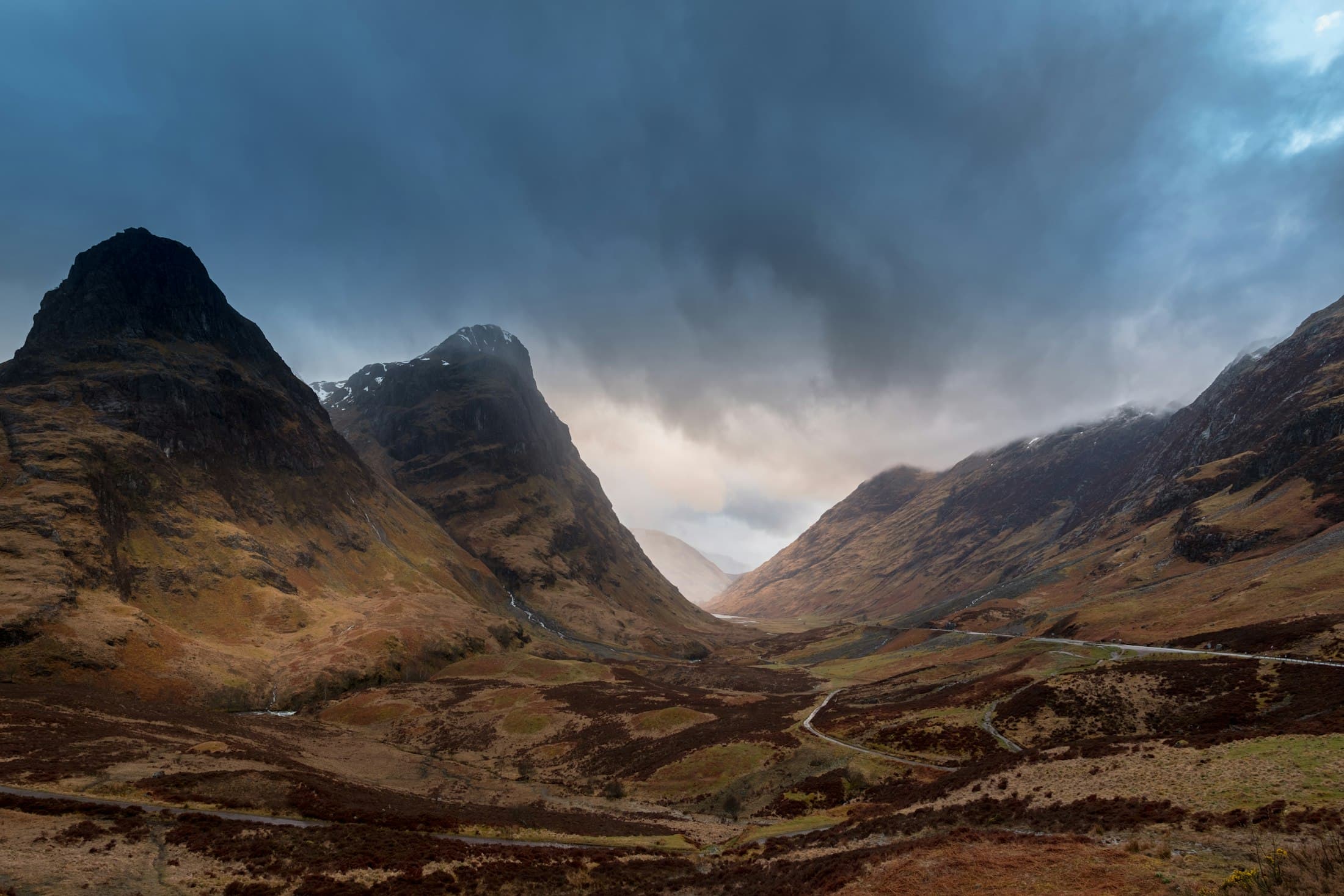 Steile, rostbraune Berge zu beiden Seiten von Glen Coe, etwas Schnee auf den Gipfeln und die Straße, die sich unter dunklem, stürmischem Himmel durchs Tal windet
