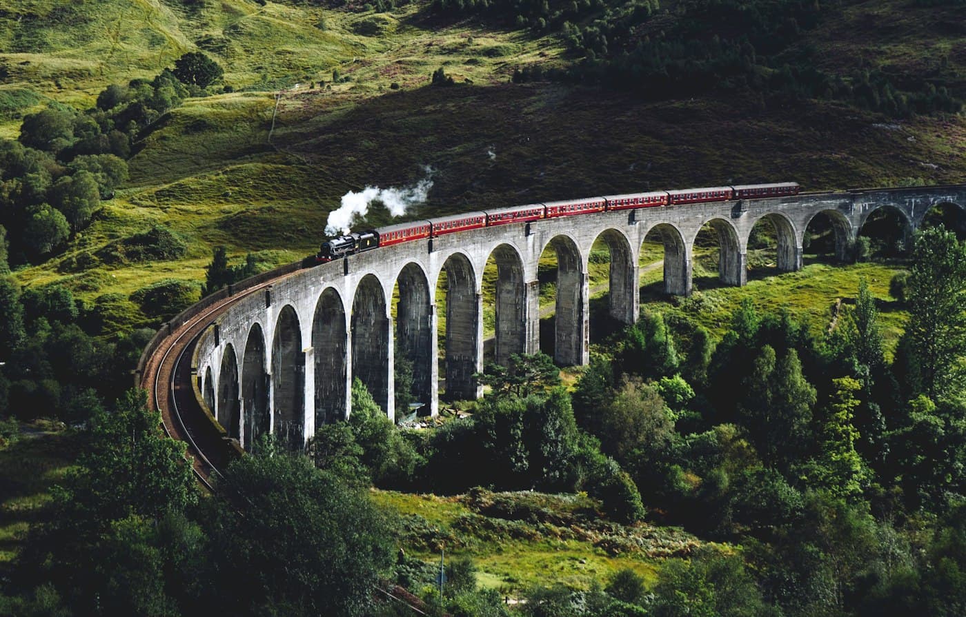 Un tren de vapor con vagones granates cruzando los arcos de piedra en curva del viaducto de Glenfinnan entre bosques y montes verdes