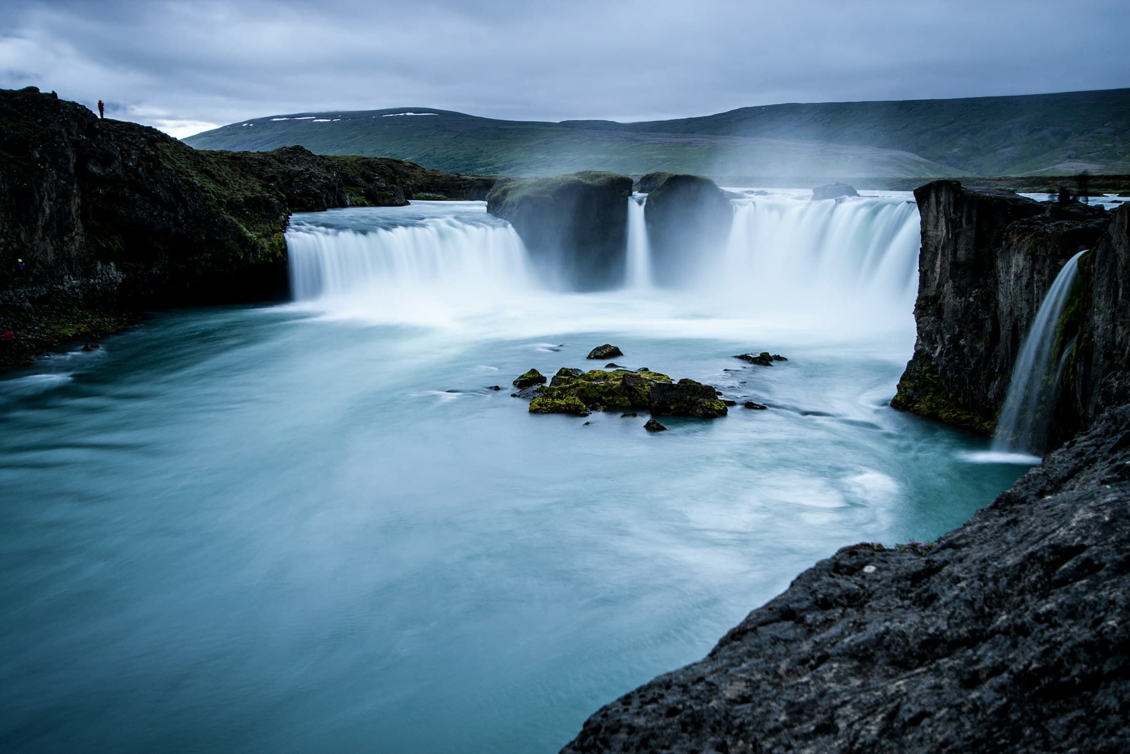 La ancha cortina de Goðafoss cayendo en una poza turquesa, entre acantilados oscuros de basalto y colinas verdes bajo un cielo gris