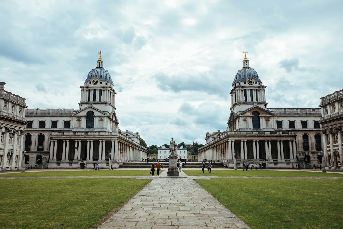 Les deux dômes de l’Old Royal Naval College se faisant face de part et d’autre d’une place gazonnée, sous un ciel nuageux, une statue sur l’allée de pierre entre eux, quelques visiteurs et, au fond, la Queen’s House blanche