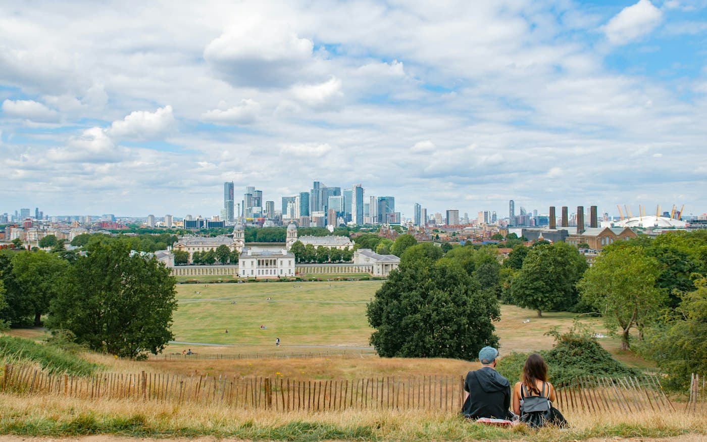 Un couple vu de dos, assis dans l’herbe sèche de la colline de Greenwich Park, qui regarde par-delà les pelouses la Queen’s House, les deux dômes de l’Old Royal Naval College et les tours de Canary Wharf