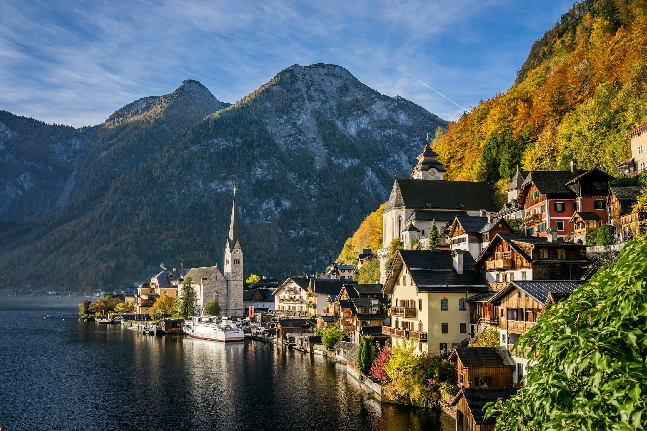 Der Kirchturm und die Holzhäuser von Hallstatt am Seeufer unter den Bergen, die Hänge herbstlich golden