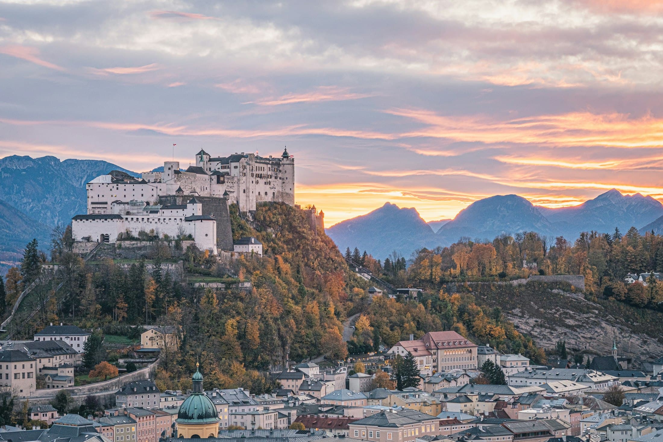 Die Festung Hohensalzburg über der Salzburger Altstadt im Abendrot, mit Herbstbäumen am Festungsberg und Bergen dahinter