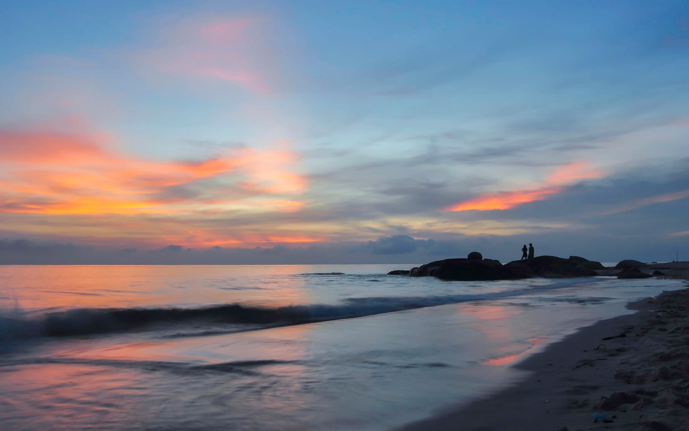 Sonnenaufgang über dem Golf von Thailand vom Strand von Hua Hin aus: orangefarbene Wolkenstreifen an einem violetten Himmel, eine Welle bricht am Ufer, und auf den dunklen Felsen am Wasser stehen zwei Menschen im Gegenlicht