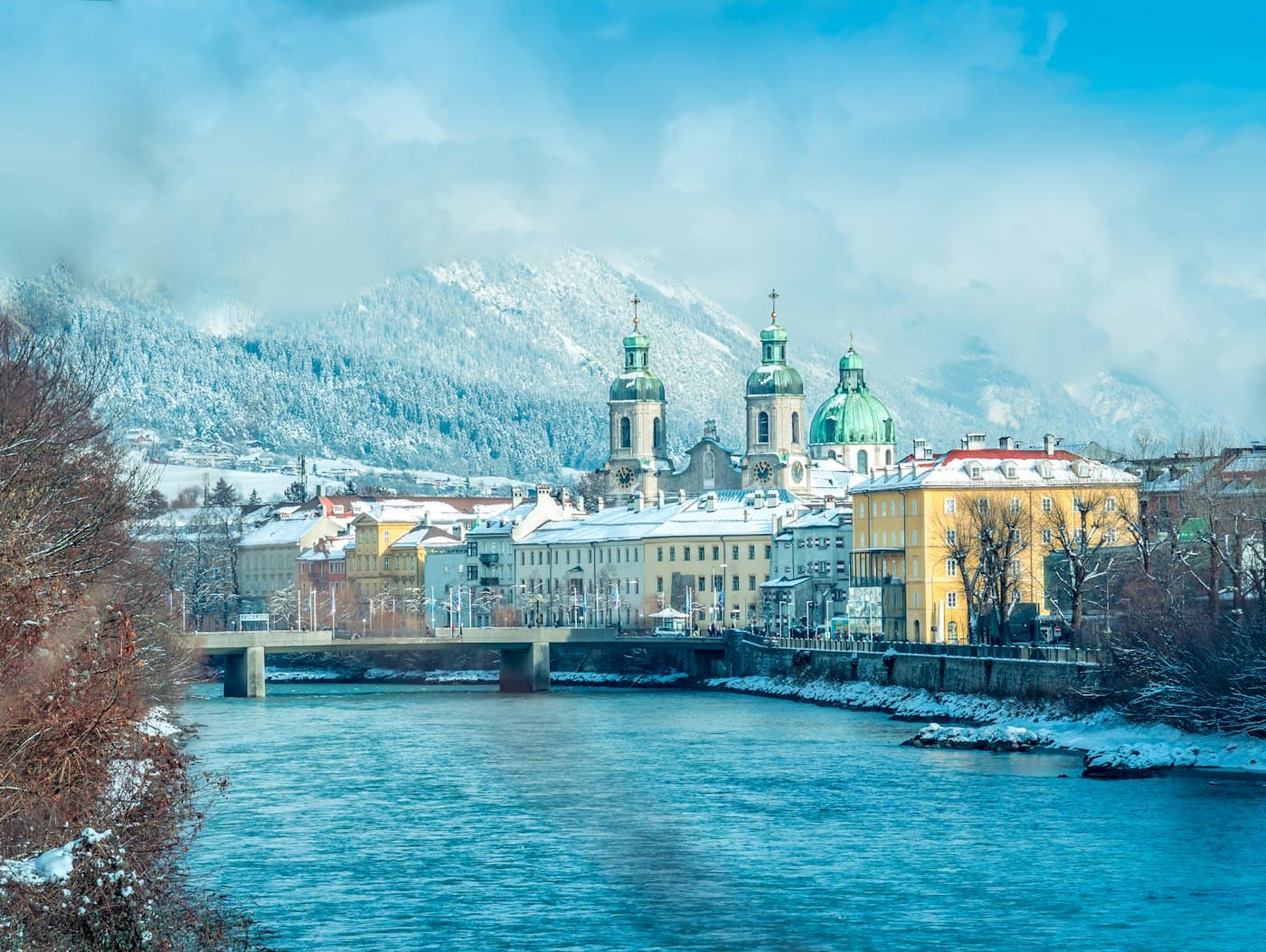 Innsbruck’s cathedral and riverside houses under fresh snow, seen across the turquoise Inn