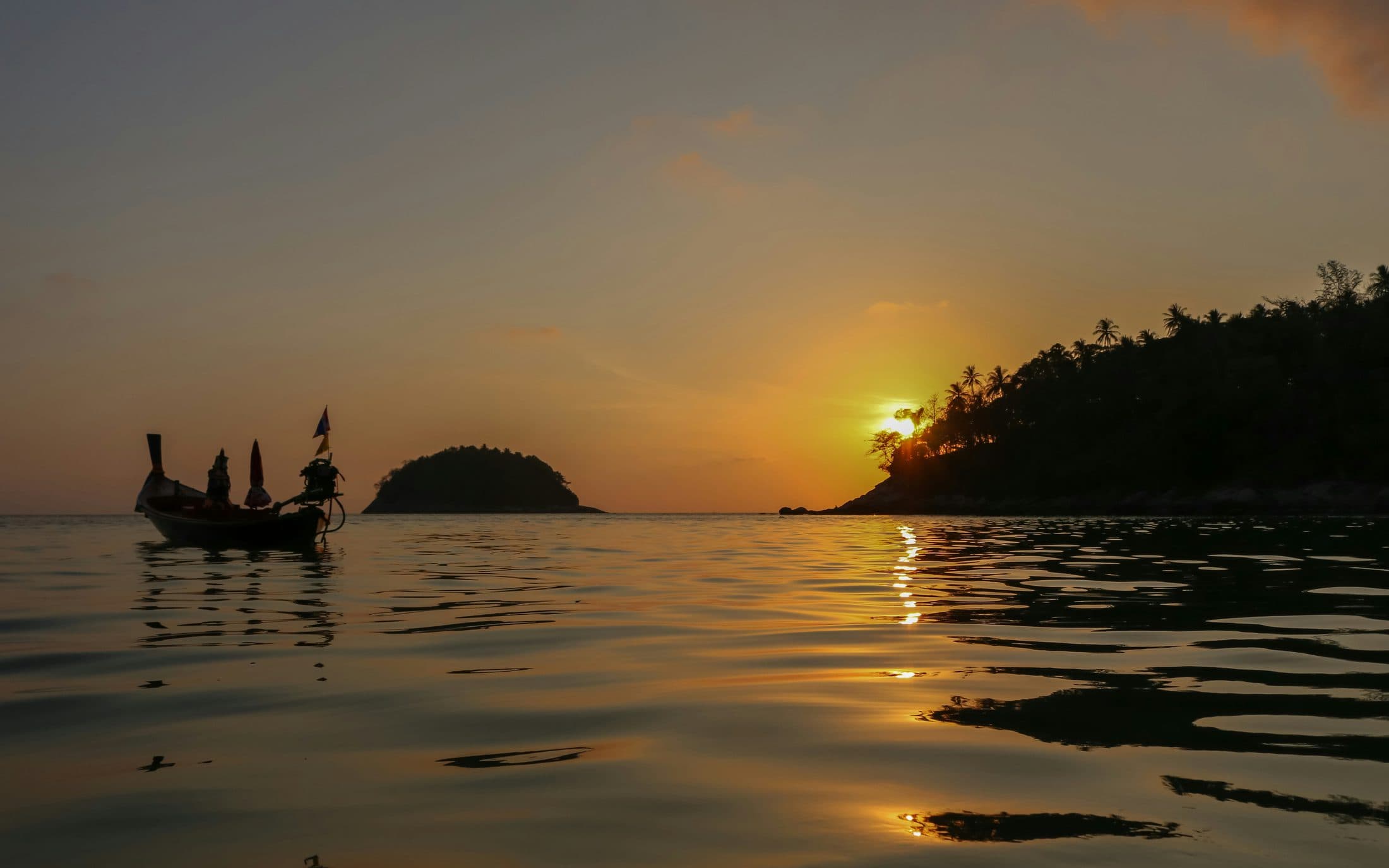 Ein Longtail-Boot als Silhouette auf der ruhigen See vor dem Strand von Kata bei Sonnenuntergang, eine kleine bewaldete Insel am Horizont und die Sonne, die hinter einer Landzunge mit Palmen versinkt und sich golden im Wasser spiegelt