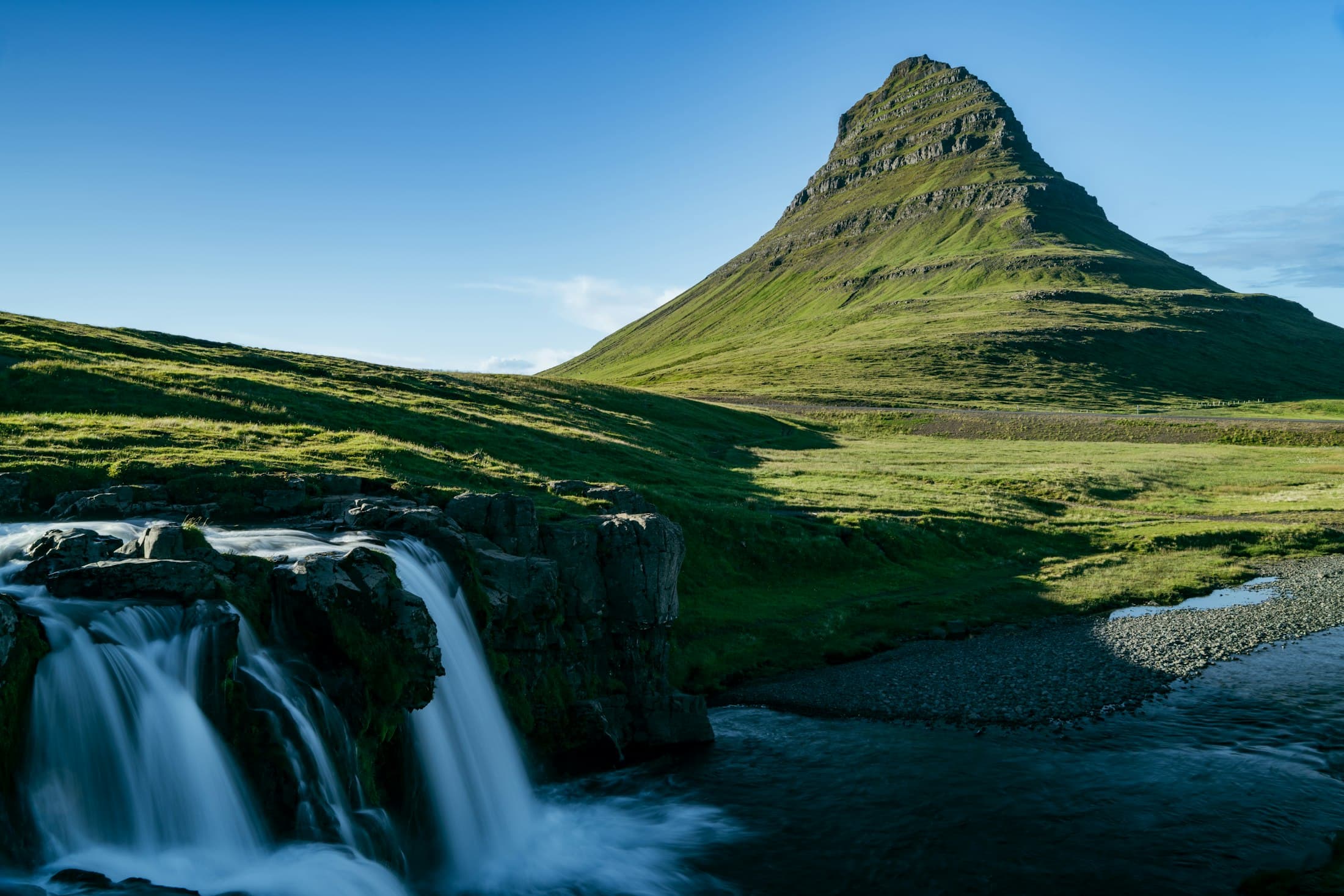 El cono verde y escalonado del Kirkjufell bajo un cielo azul, con los dos saltos de Kirkjufellsfoss cayendo sobre roca oscura en primer plano