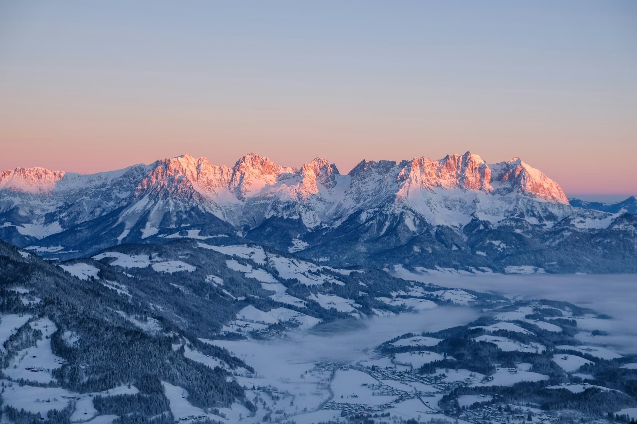 Verschneite Kalkgipfel im rosa Alpenglühen über einem nebligen Wintertal bei Kitzbühel