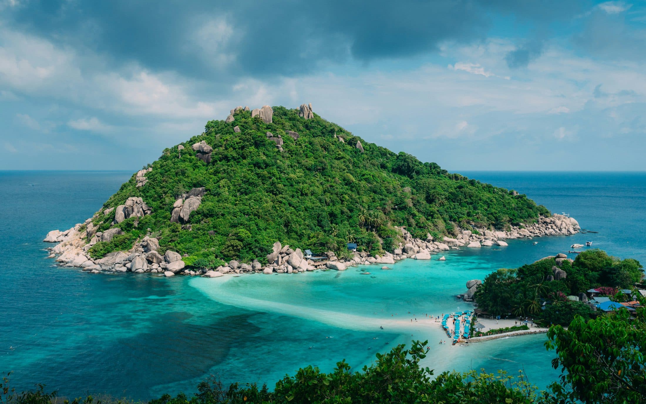 Eine steile grüne Insel mit Granitblöcken auf dem Gipfel, über türkisfarbenes Flachwasser durch eine geschwungene weiße Sandbank mit einer kleineren bewaldeten Insel mit Bungalows, Sonnenschirmen und ein paar Badenden verbunden, Koh Nang Yuan, unter bewölktem Himmel