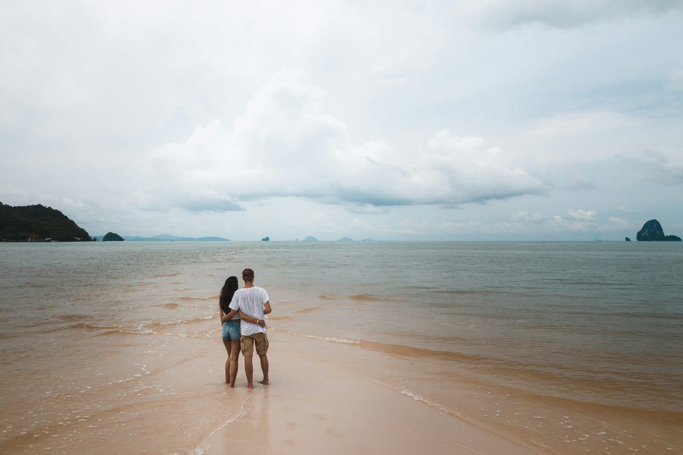 Una coppia vista di spalle, abbracciata e a piedi nudi su un banco di sabbia chiara nelle isole Koh Yao, che guarda un mare calmo punteggiato di lontane isole calcaree sotto un cielo nuvoloso