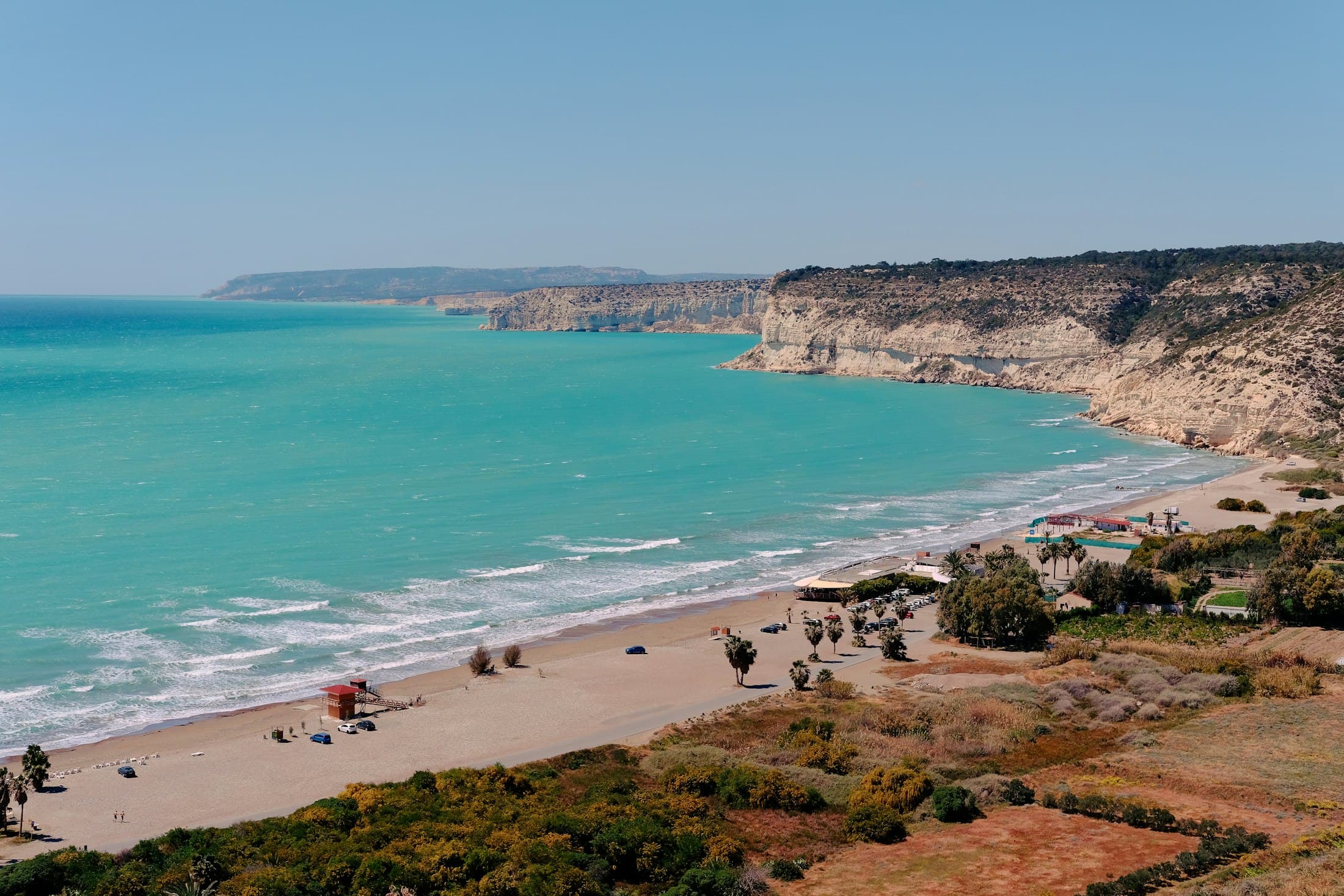 The long beach below Kourion seen from above, turquoise water breaking white on the sand and pale cliffs running west to a distant headland
