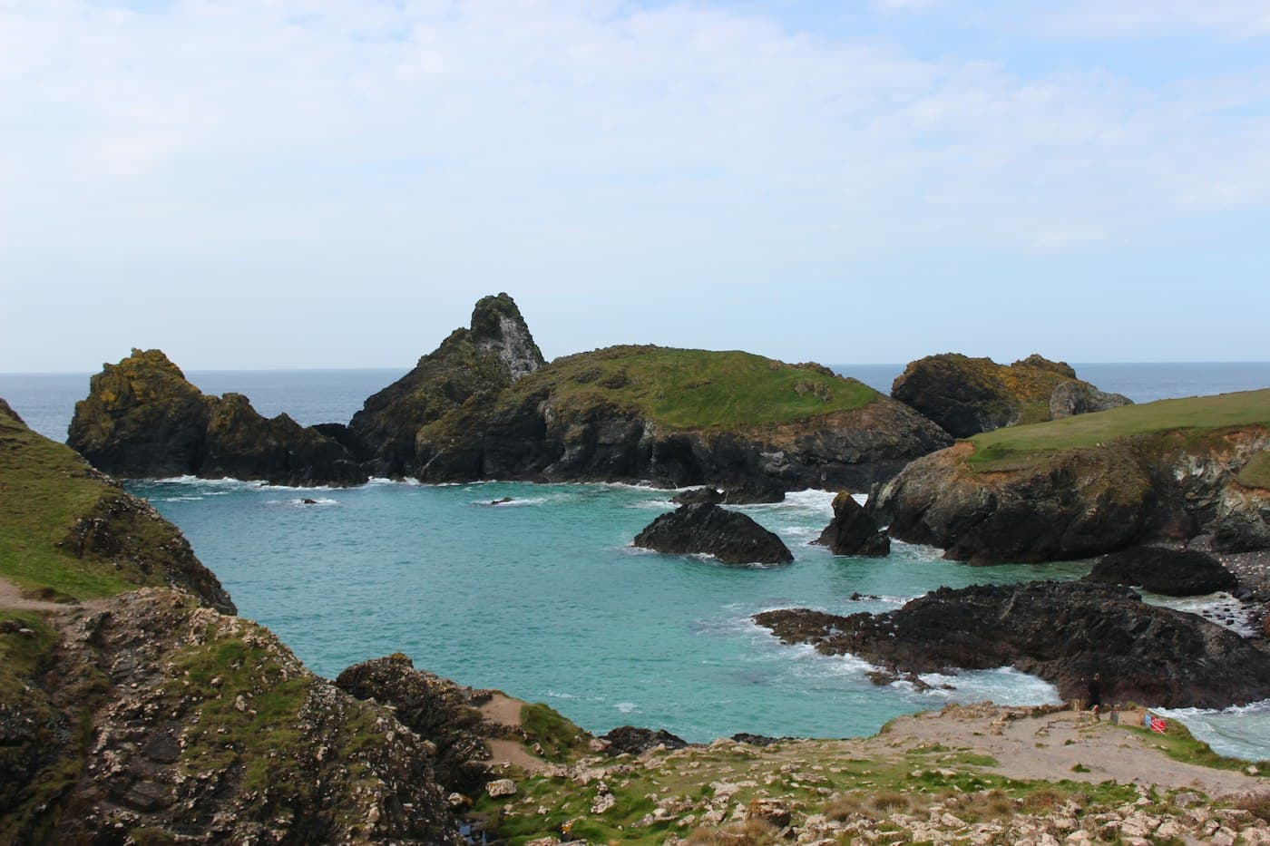 I faraglioni e l’isola erbosa di Kynance Cove che si alzano dall’acqua turchese, visti dai pendii sopra la caletta, sul Lizard