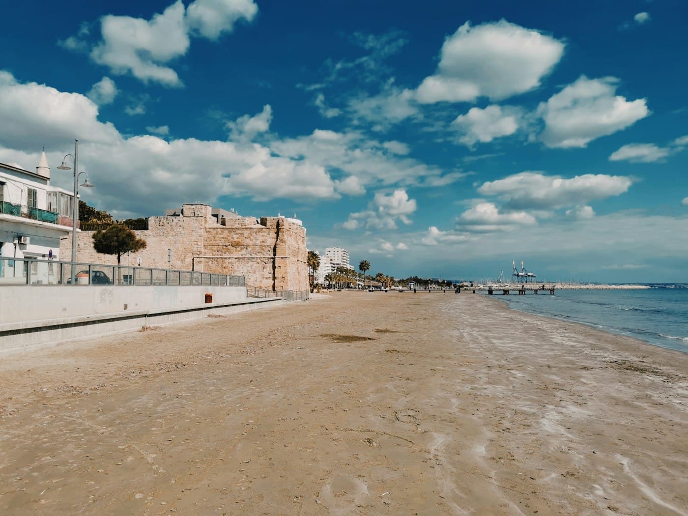 The beach at Finikoudes in Larnaca under a blue sky with white clouds, the stone walls of the medieval fort and the palm-lined seafront beyond