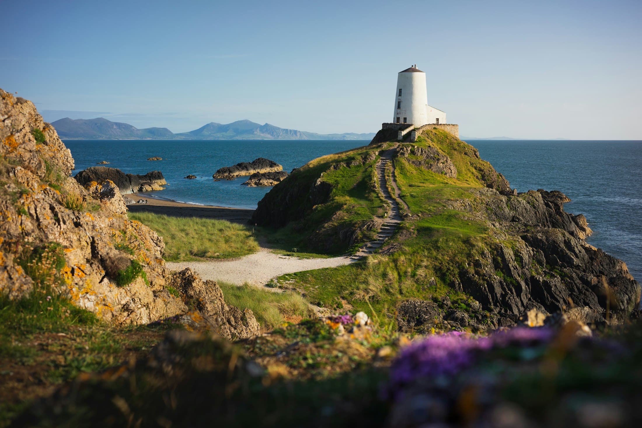 Der weiße Turm von Tŵr Mawr auf seinem grasbewachsenen Fels am Ende von Ynys Llanddwyn, Steinstufen führen von einer kleinen Sandbucht hinauf, dunkle Felsen im blauen Meer und Berge jenseits des Wassers