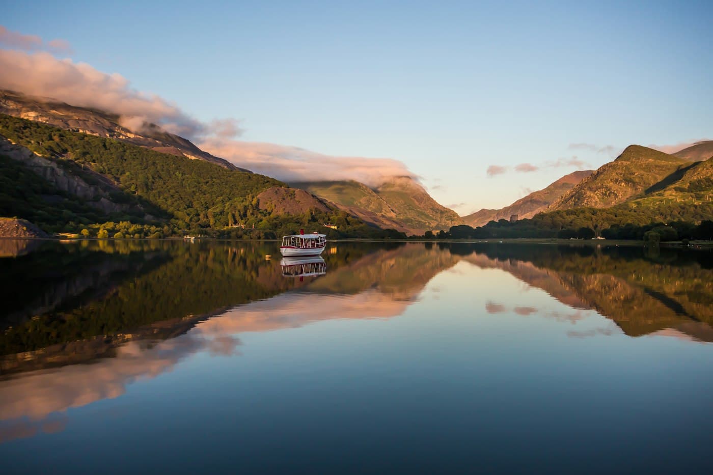 A small white boat on the still water of Llyn Padarn, the mountains of Eryri lit by a low sun and mirrored in the lake, with the tower of Dolbadarn Castle small among the trees on the far shore