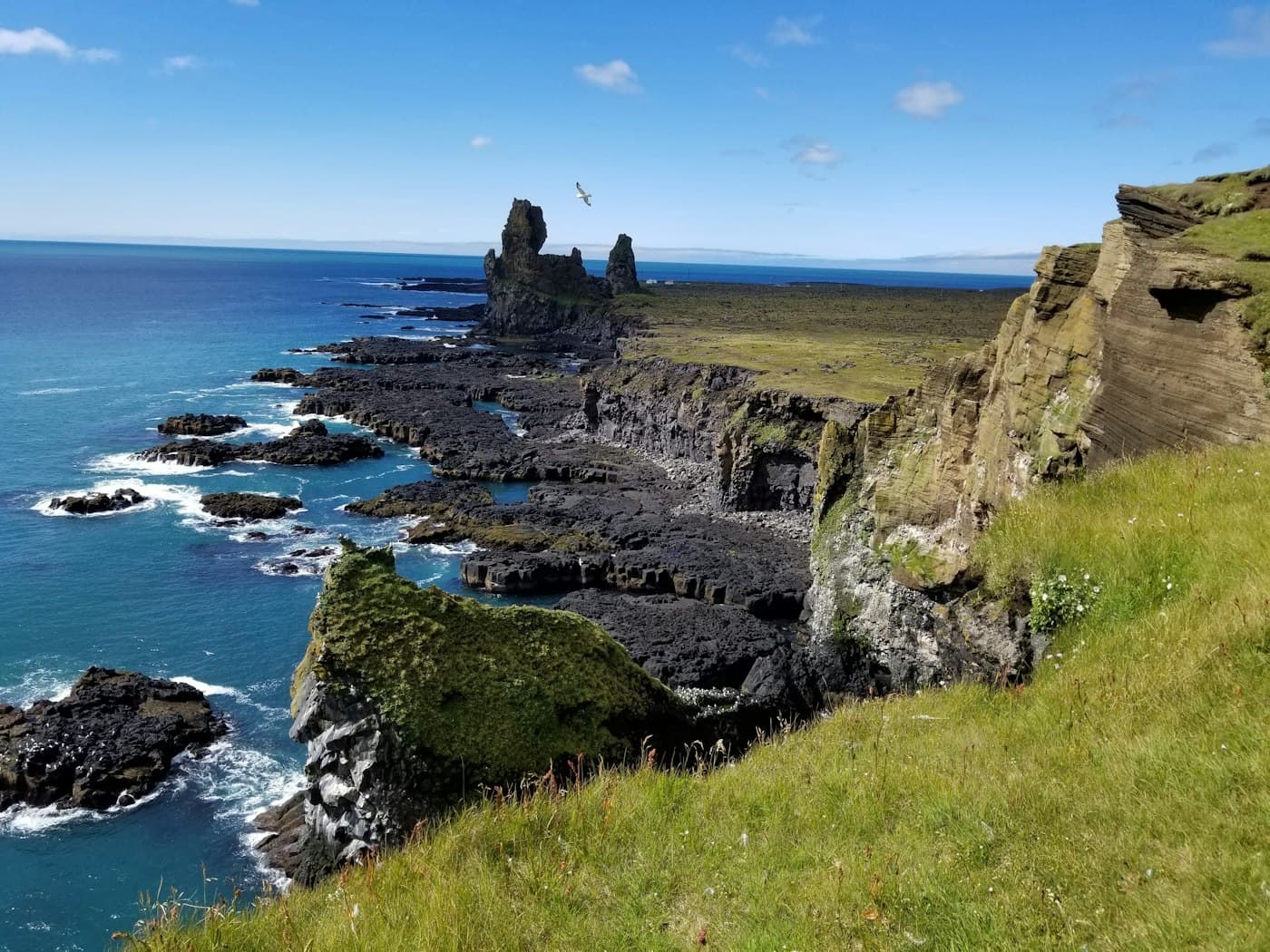 Los farallones de Lóndrangar tras una línea de acantilados de basalto y arrecifes negros, vistos desde lo alto de un acantilado de hierba en un día de sol