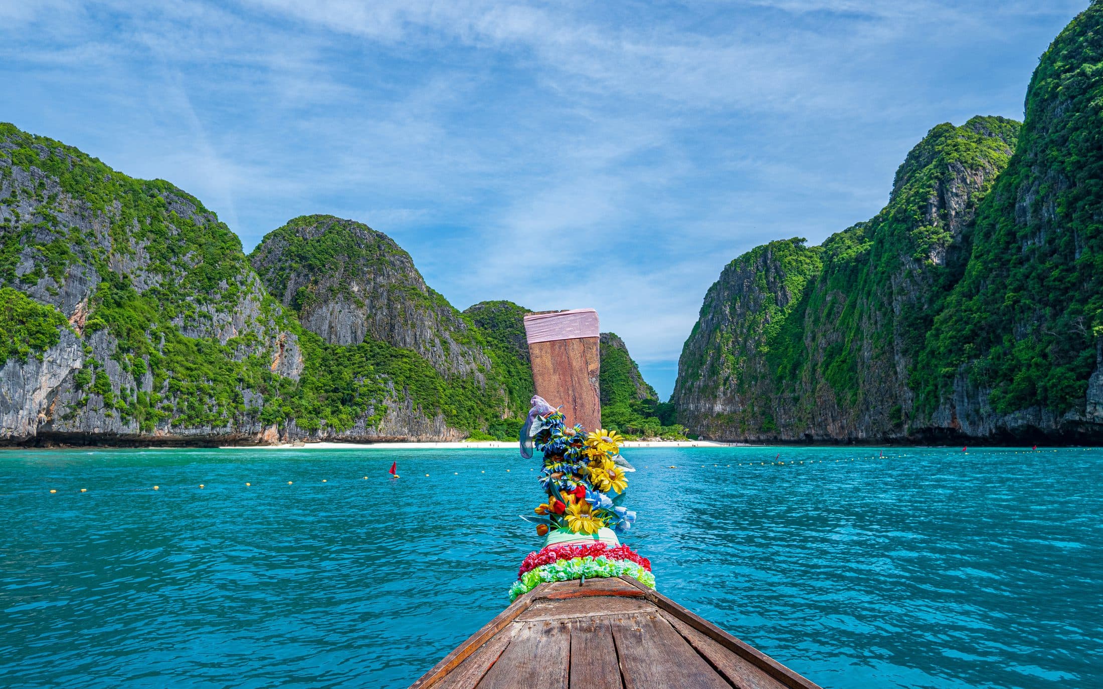 Die Maya Bay auf Koh Phi Phi Leh, gesehen vom Bug eines mit Blumengirlanden geschmückten Longtail-Boots: türkisfarbenes Wasser, eine Bojenkette quer durch die Bucht und ein Streifen weißer Sand zwischen steilen, grünen Kalksteinfelsen