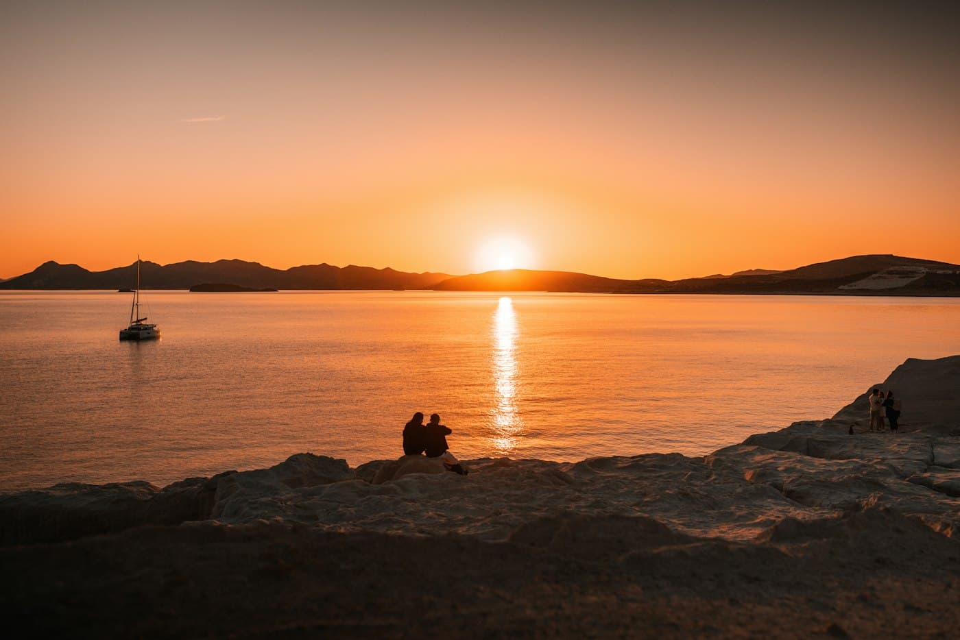 A couple sitting on the rocks of Milos as the sun sets over the bay, a sailing boat at anchor