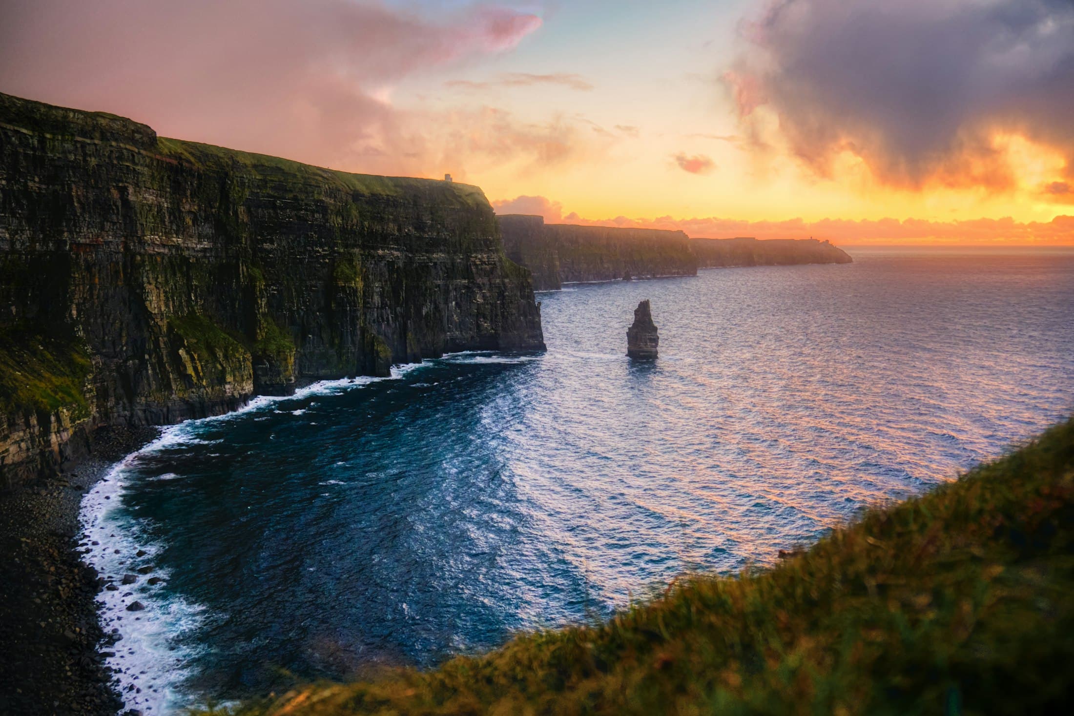 Le Scogliere di Moher al tramonto: roccia scura a strati che precipita su una riva curva, un faraglione solitario e la torre di O’Brien in cima, sotto un cielo arancione