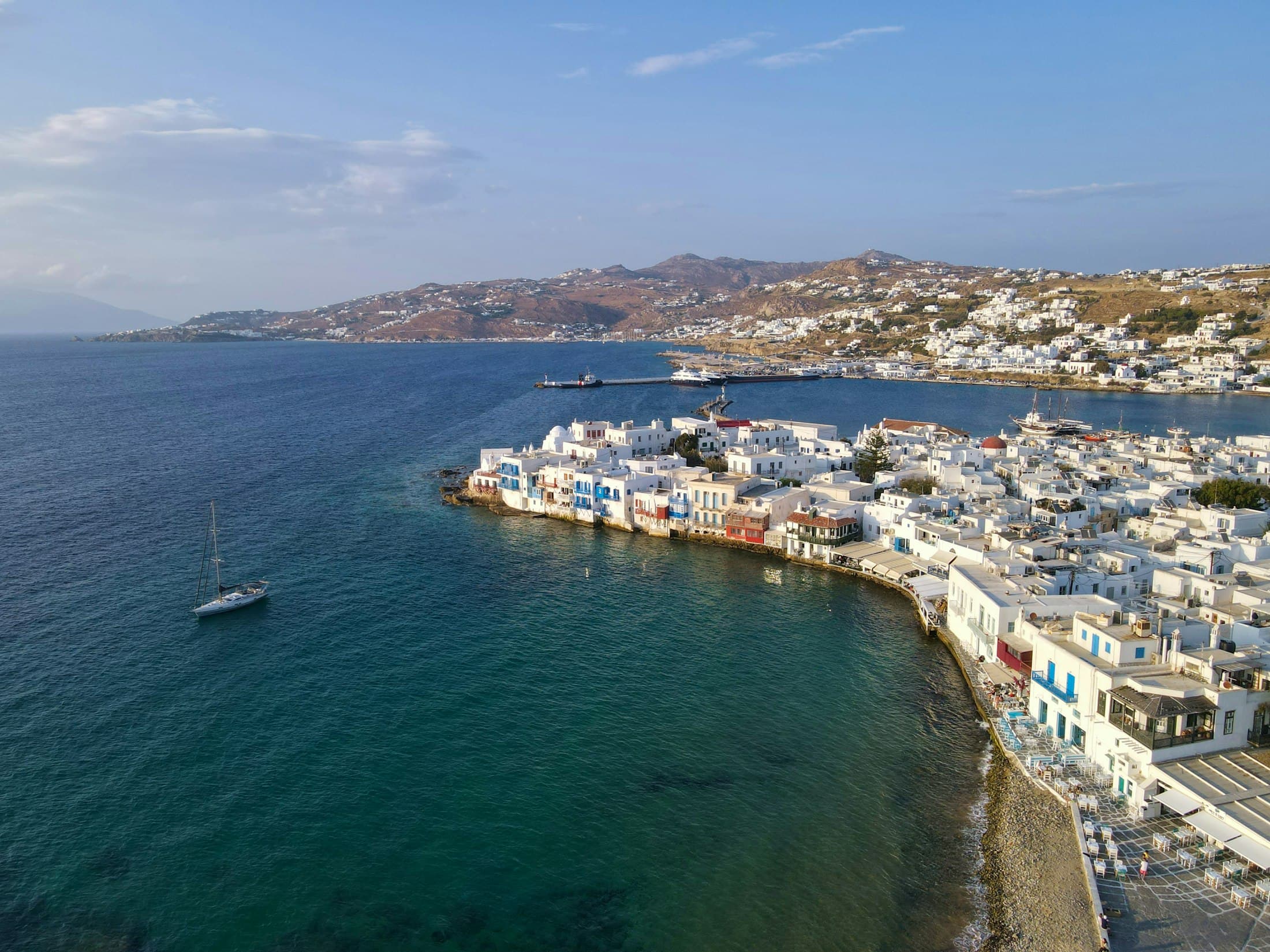 Klein-Venedig und der weiße Hauptort von Mykonos von oben, mit einem Segelboot in der Bucht und den Hügeln der Insel dahinter