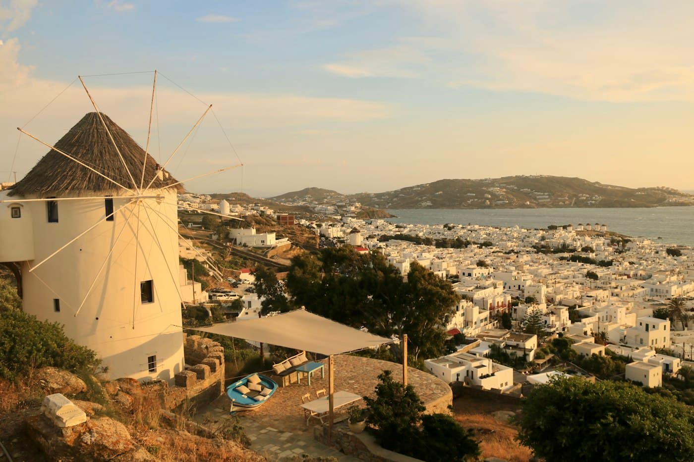 Eine Windmühle über dem weißen Hauptort von Mykonos im Abendlicht, dahinter die Bucht und die Hügel