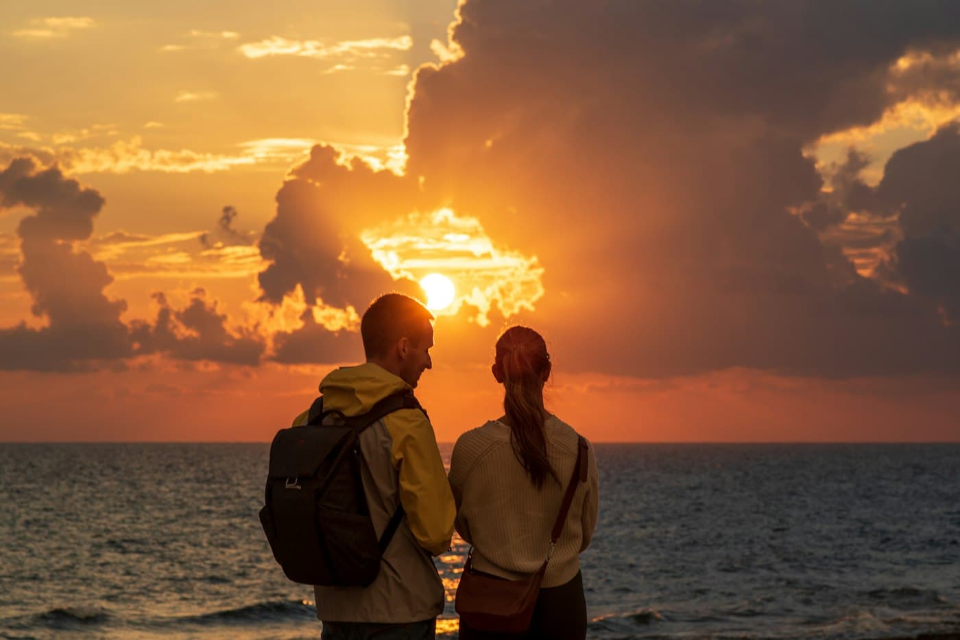 A couple seen from behind on the shore at Paphos, the sun setting into the sea through a break in dark clouds