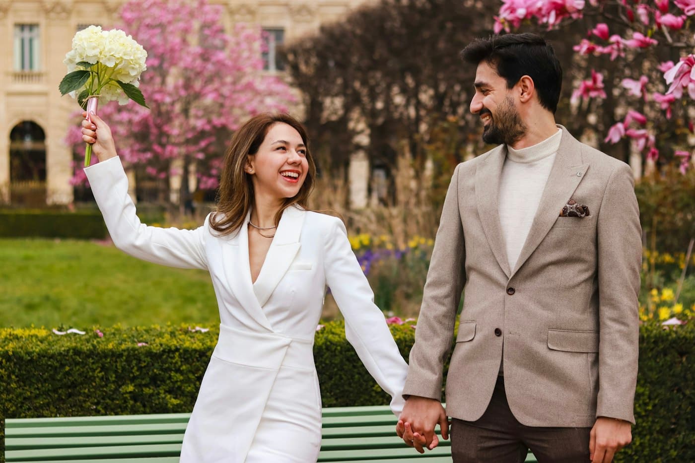 A bride in a white suit raising her bouquet, hand in hand with her groom in a Paris garden