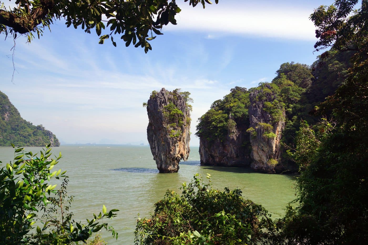 Khao Tapu, uno snello pinnacolo di calcare che si alza dall’acqua verde della baia di Phang Nga, con pareti calcaree boscose alla sua destra e le foglie e i rami degli alberi sulla riva a incorniciare la vista