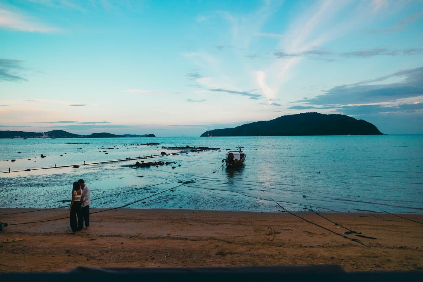 Ein Paar eng umschlungen an einem Strand auf Phuket in der Abenddämmerung, bei Ebbe, ein Longtail-Boot im flachen Wasser vertäut, Leinen auf dem nassen Sand und eine bewaldete Insel jenseits der Bucht unter blassblauem Himmel