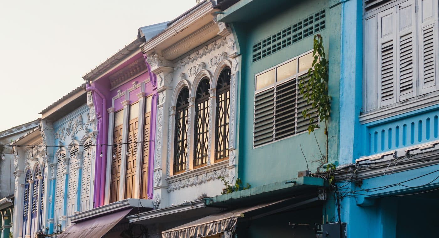 Restaurierte Ladenhäuser in der Altstadt von Phuket in der späten Nachmittagssonne: hohe Bogenfenster mit weißem Stuck, Holztüren und Lamellenläden, Fassaden in Flieder, Hellblau, Weiß, Türkis und kräftigem Blau, Markisen über den Ladenfronten