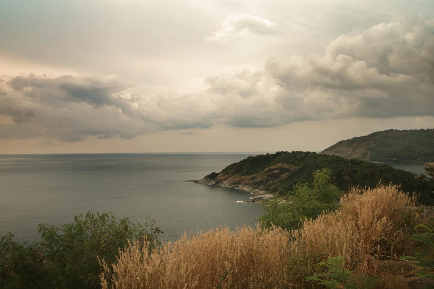 Der Blick vom Kap Promthep unter schweren Wolken: eine bewaldete Landzunge, die in ein ruhiges, graugrünes Meer hinausläuft, dahinter ein zweiter Hügel, im Vordergrund trockene goldene Gräser und Büsche