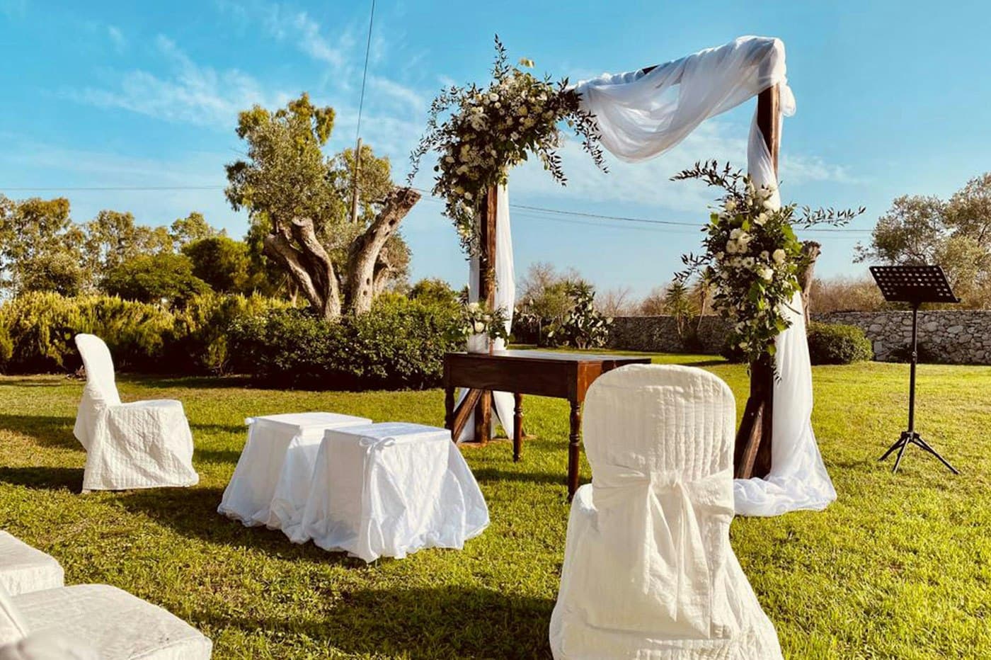 A ceremony arch with white drapes on a lawn among olive trees