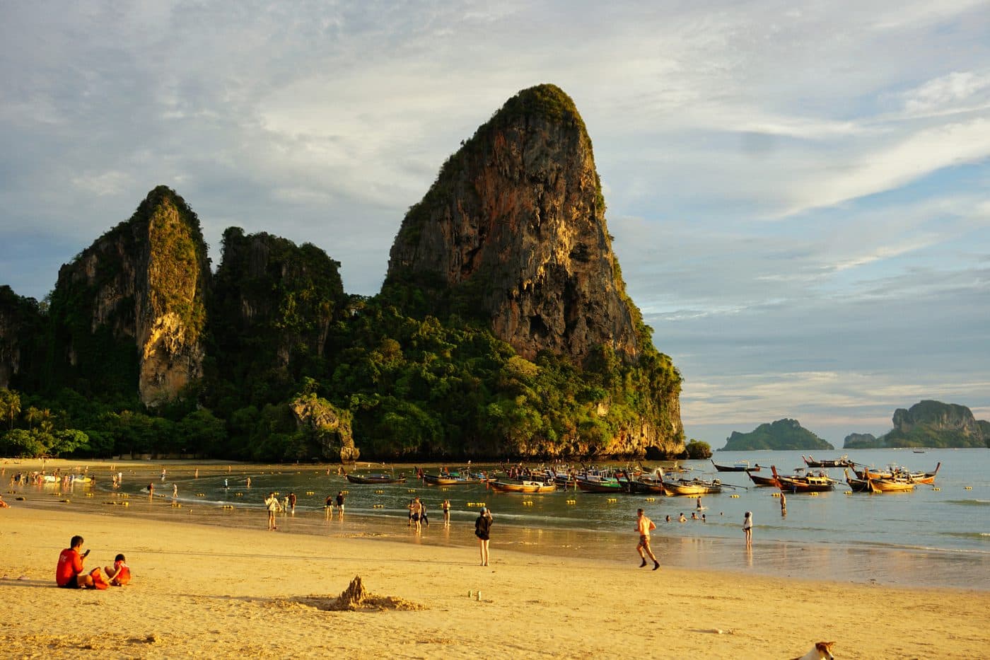 La spiaggia di Railay nella luce calda della sera: alti pinnacoli di calcare dorati sopra la giungla, barche a coda lunga all’ancora nell’acqua bassa e persone che camminano sull’ampia sabbia della bassa marea