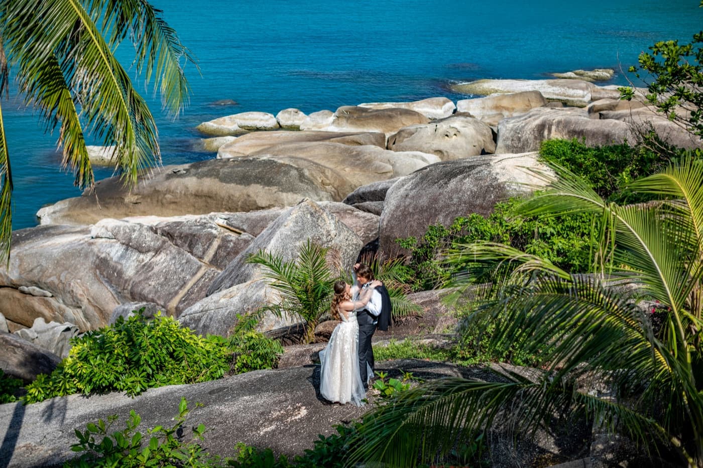 A bride in a long white dress and a groom in a dark waistcoat, his jacket over his shoulder, holding each other among great granite boulders, palms and green shrubs above a turquoise sea on Koh Samui