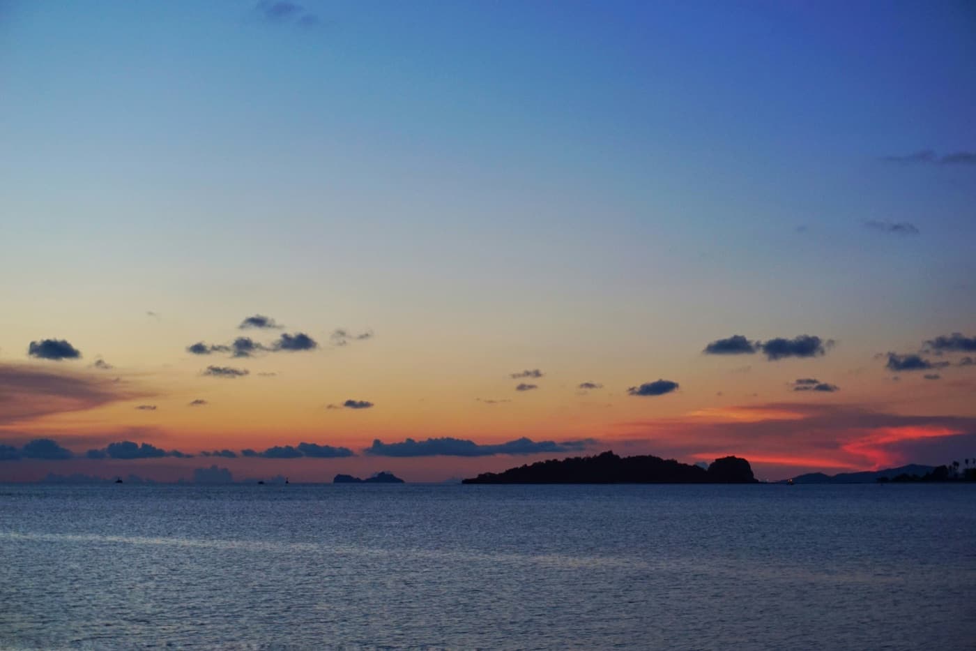 A calm sea off Koh Samui just after sunset, a low wooded island and a few small islets dark against an orange horizon, with a deep blue sky and small clouds above