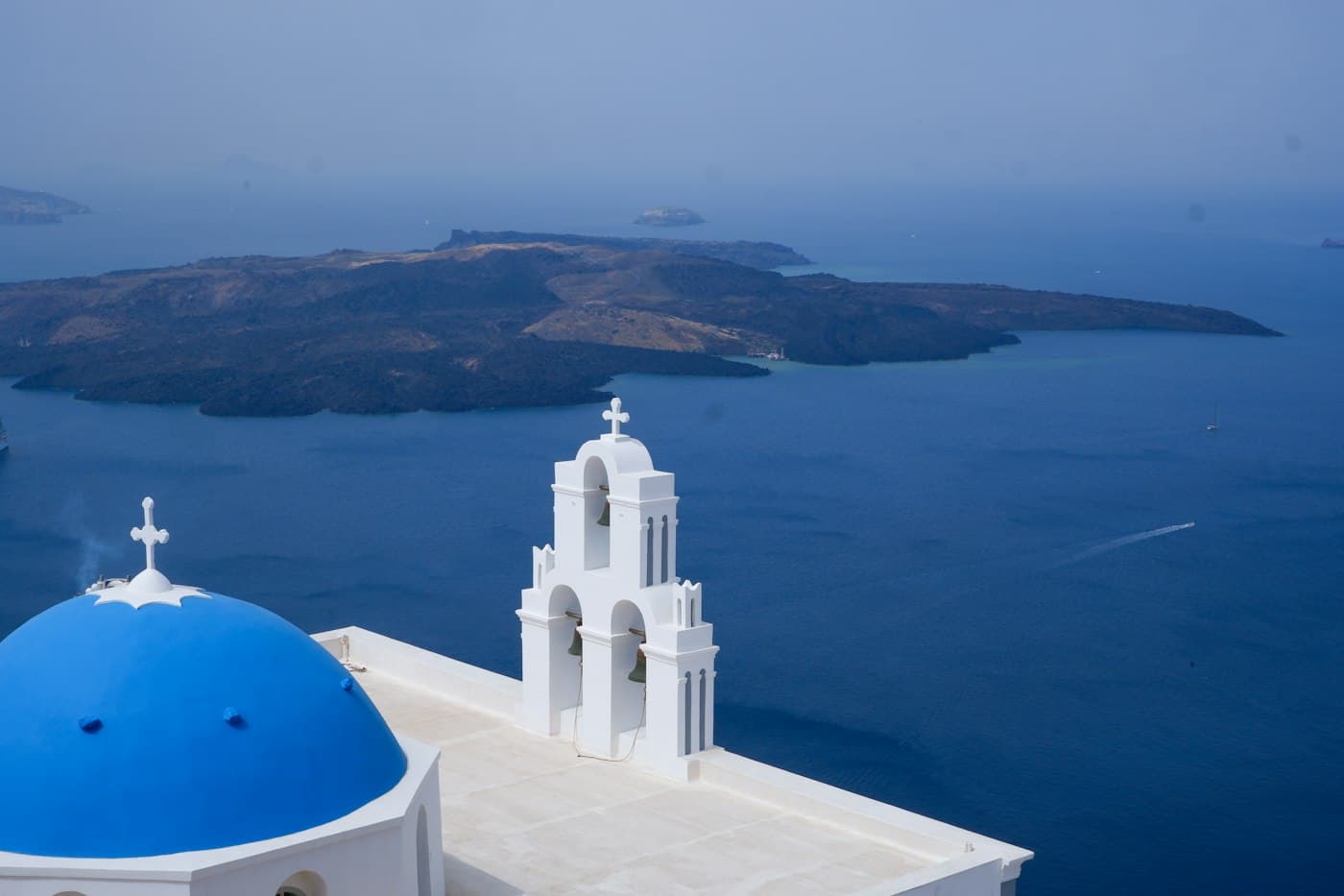 Una cúpula azul y un campanario blanco sobre la caldera de Santorini, con las islas volcánicas en medio de la bahía