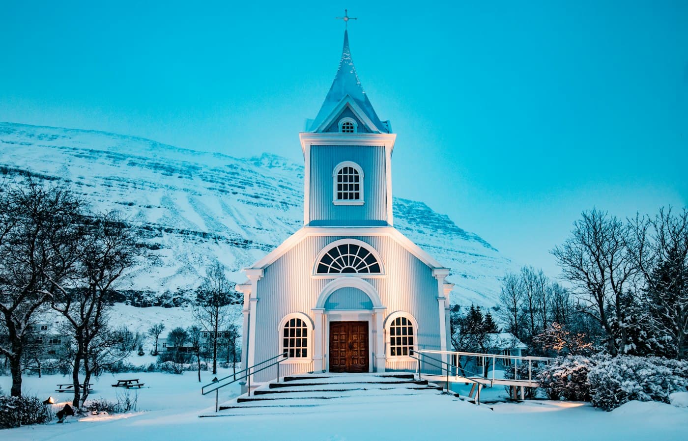 Die hellblaue Holzkirche von Seyðisfjörður im tiefen Schnee in der Dämmerung, von unten beleuchtet, dahinter ein verschneiter Berghang