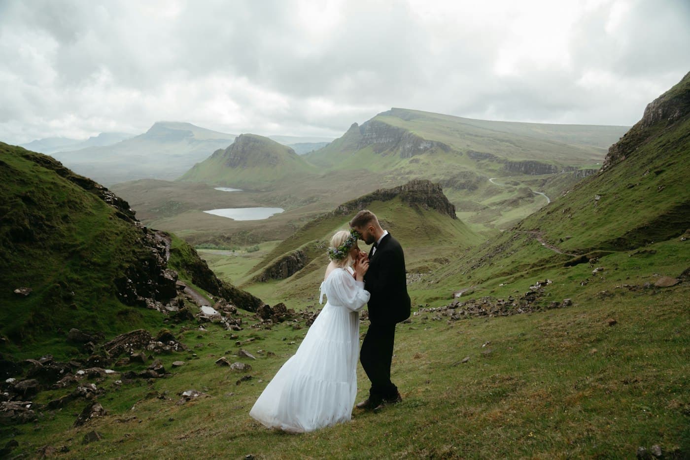 Una novia con vestido blanco largo y corona de flores y un novio con traje oscuro, frente con frente en una ladera verde del Quiraing, con sus riscos, una pequeña laguna y los montes de Skye detrás, bajo nubes grises