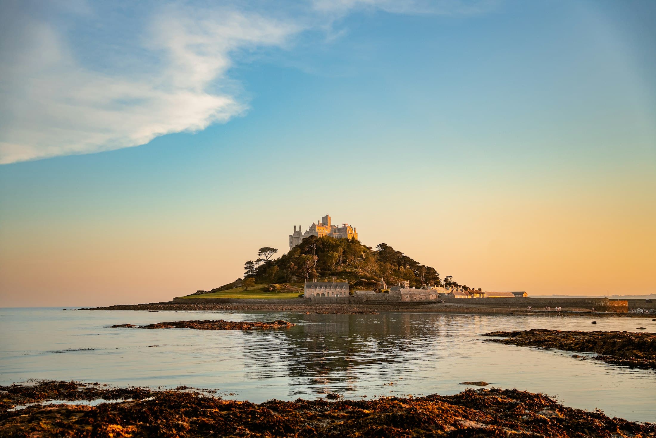 St Michael’s Mount mit seiner Burg in der tiefen Abendsonne, gespiegelt im ruhigen Wasser vor Marazion, im Vordergrund tangbedeckte Felsen und auf dem Damm ein paar winzige Menschen