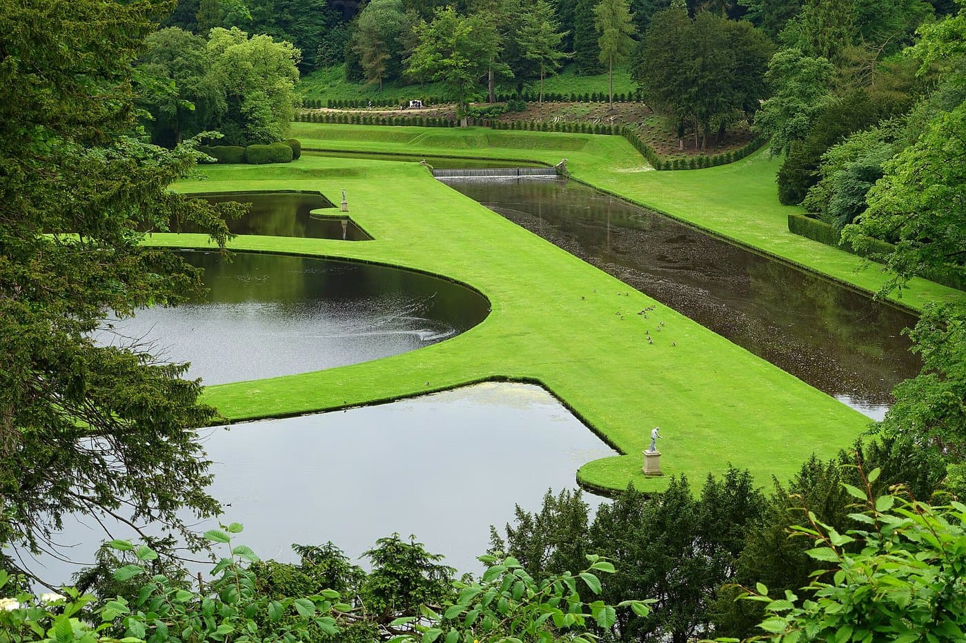 Die geschwungenen Teiche und der Kanal des Wassergartens von Studley Royal zwischen gepflegten Rasenflächen mit einer Statue, von Bäumen gerahmt und vom Hang darüber gesehen